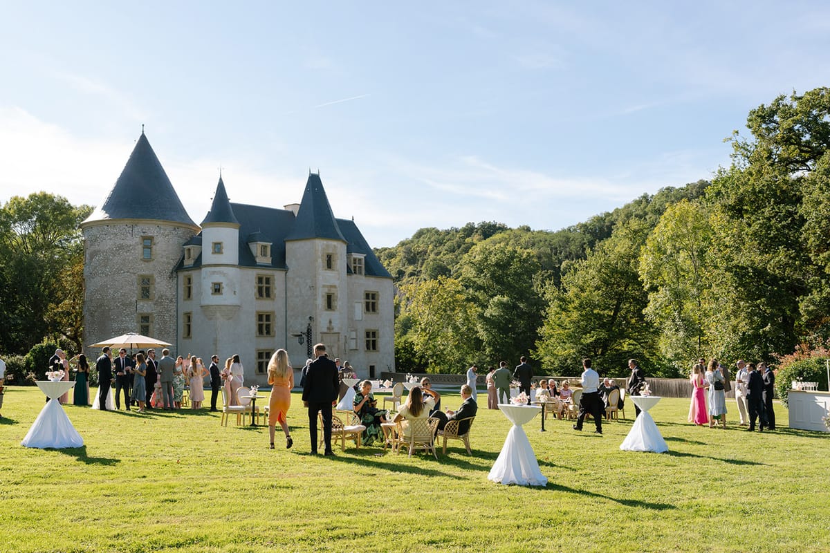 Forty guests on chateau lawn cocktail hour with white tables, rattan chairs, and conical tower behind