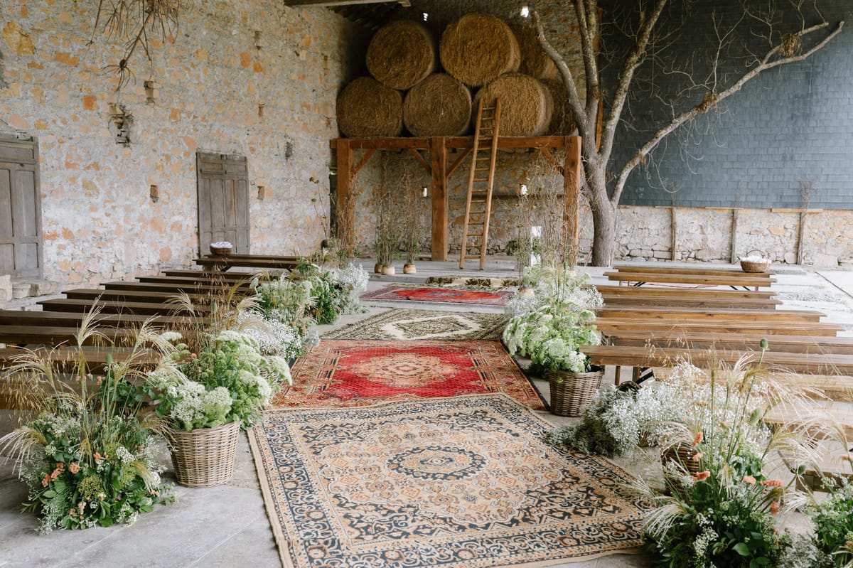 Rustic stone barn ceremony setup with vintage rugs lining the aisle, hay bale altar, and meadow-style floral baskets