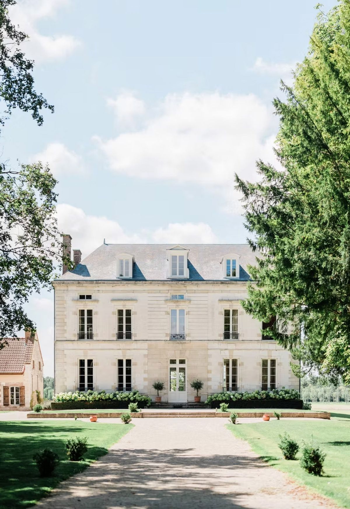 Cream stone French chateau with slate mansard roof, wrought iron balconies, white hydrangeas, and gravel driveway