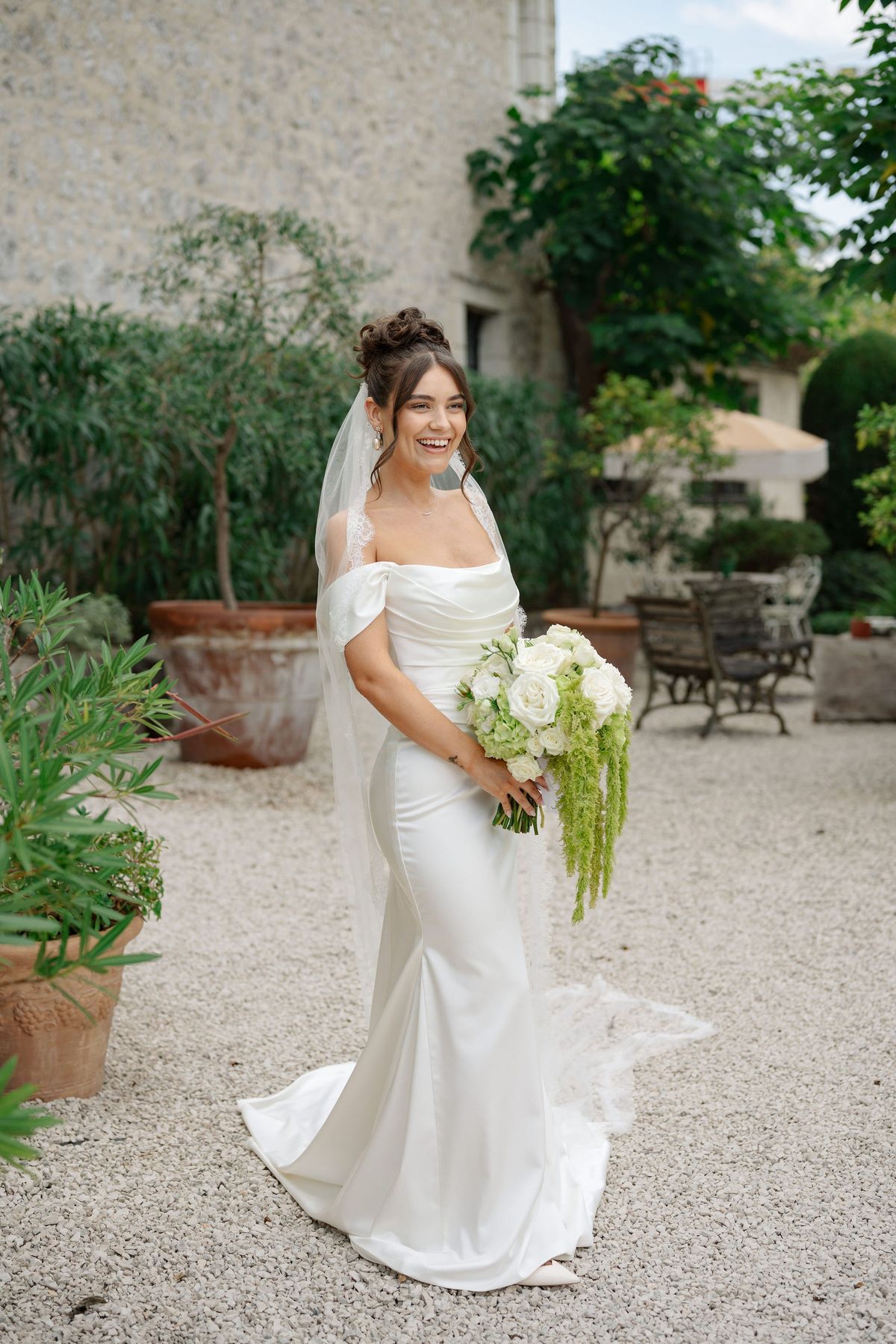 Bride in silk off-shoulder gown holding white bouquet in venue courtyard