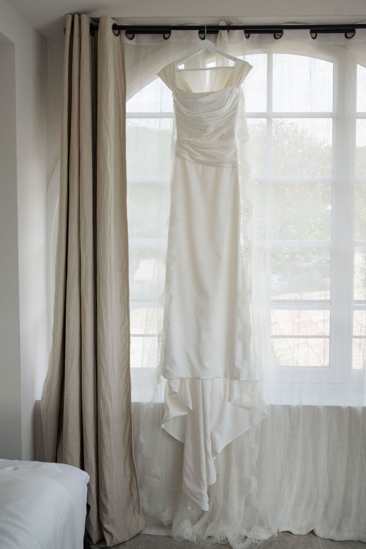 Wedding dress hanging in arched window with linen curtains and lace train