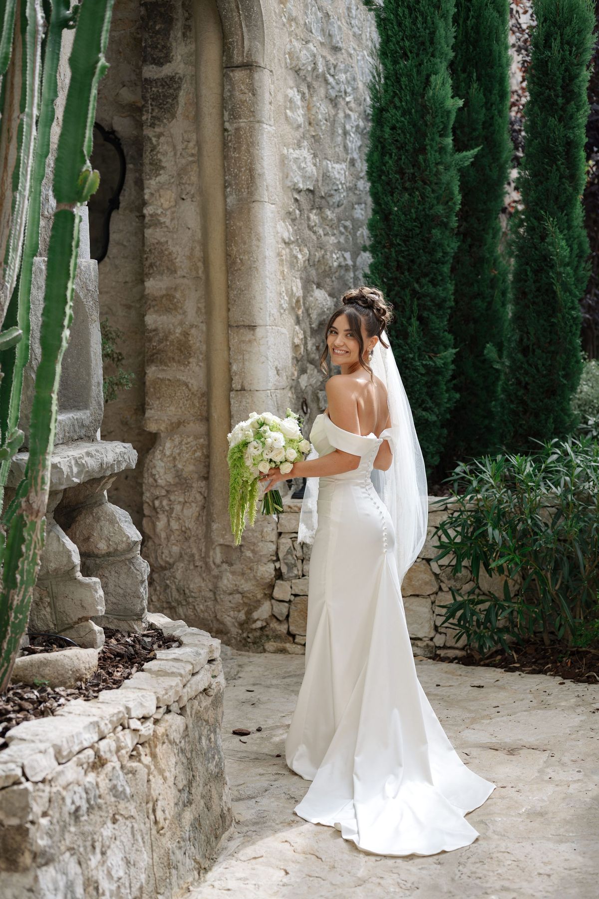 Bride looking over shoulder beside stone archway and cypress trees