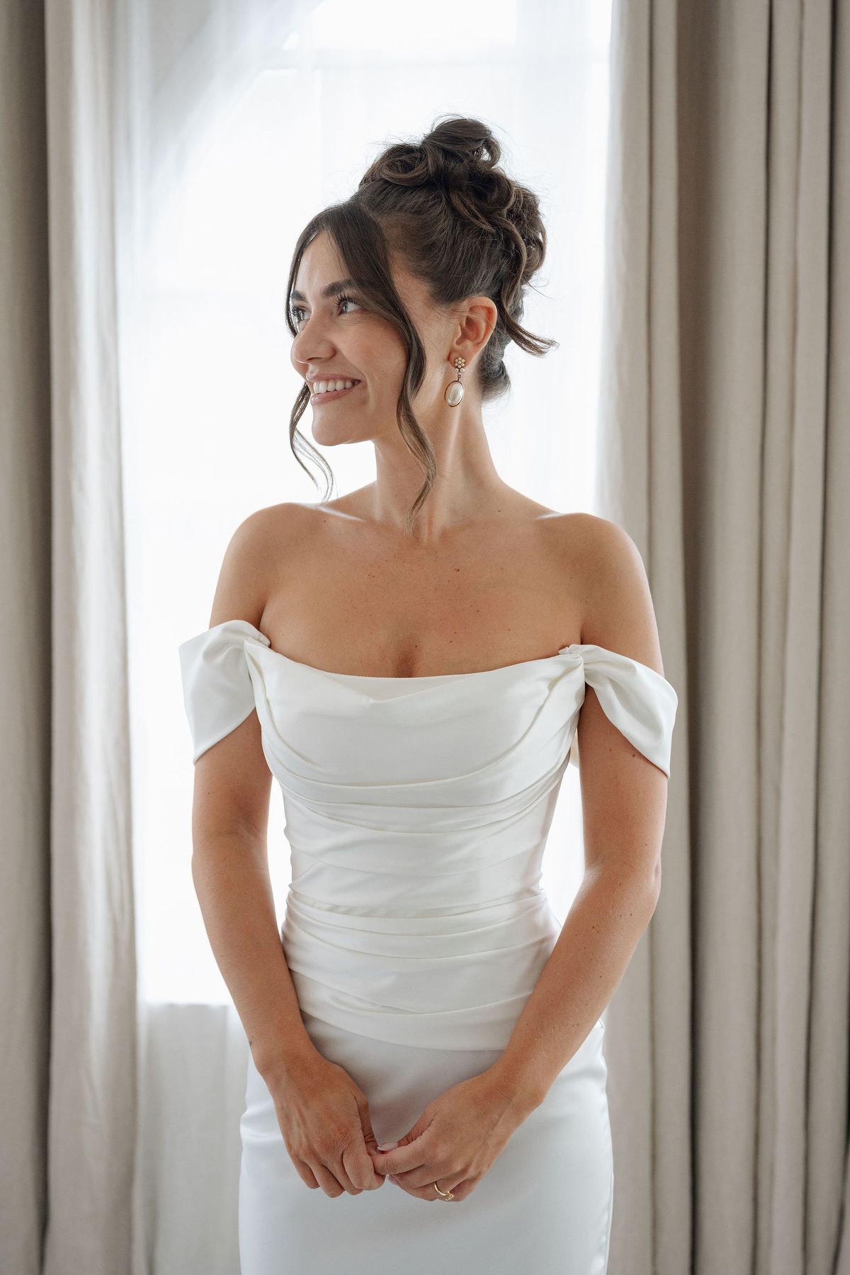 Bride in off-shoulder satin gown with chignon by sheer curtained window