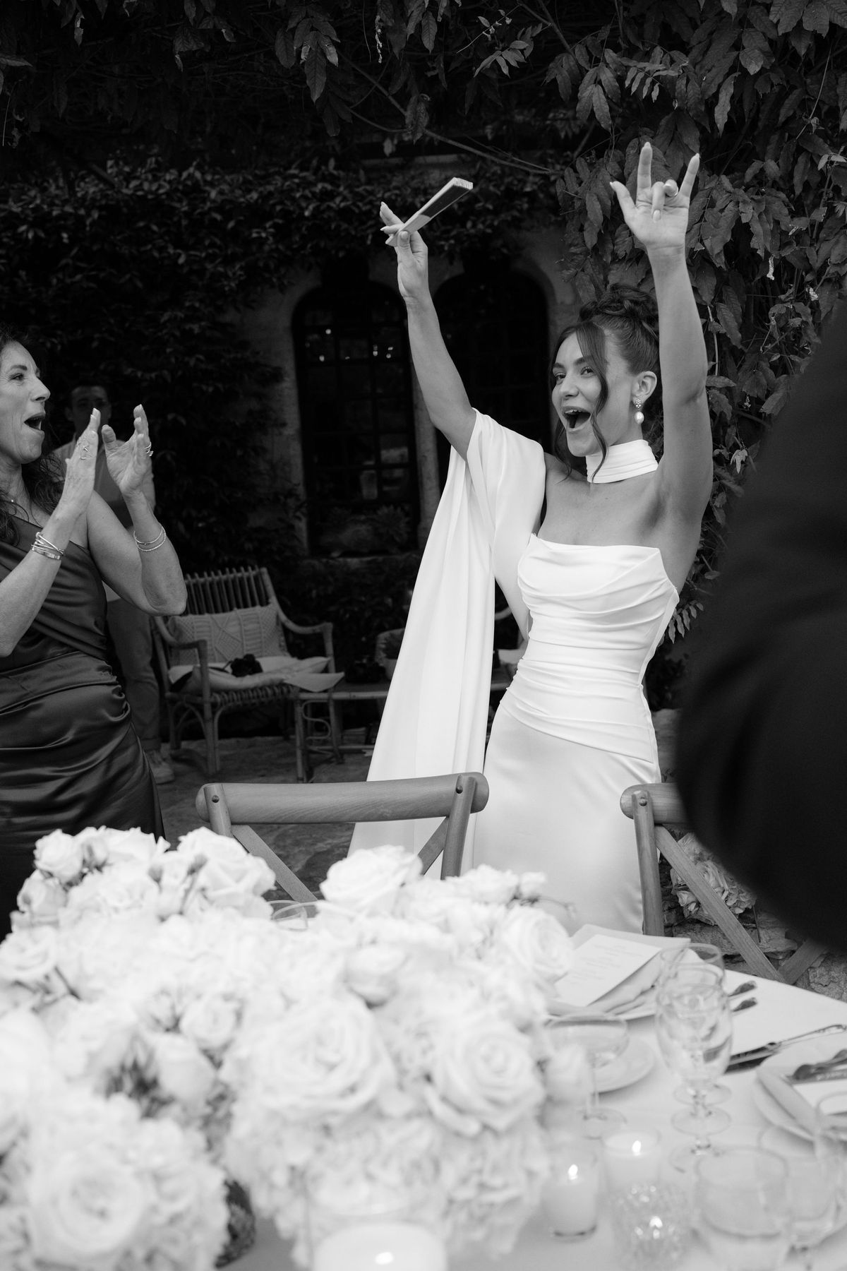 Bride celebrating with arms raised at reception table in black and white