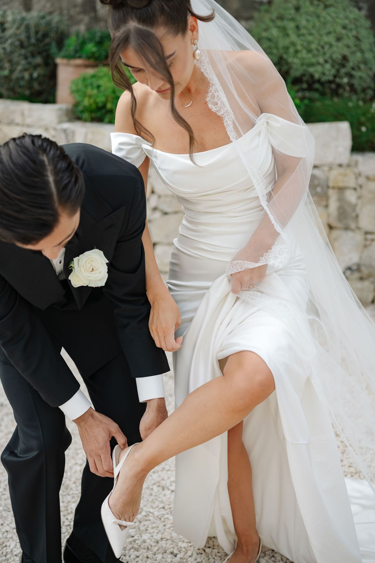 Groom kneeling to help bride adjust her shoe in garden during portraits