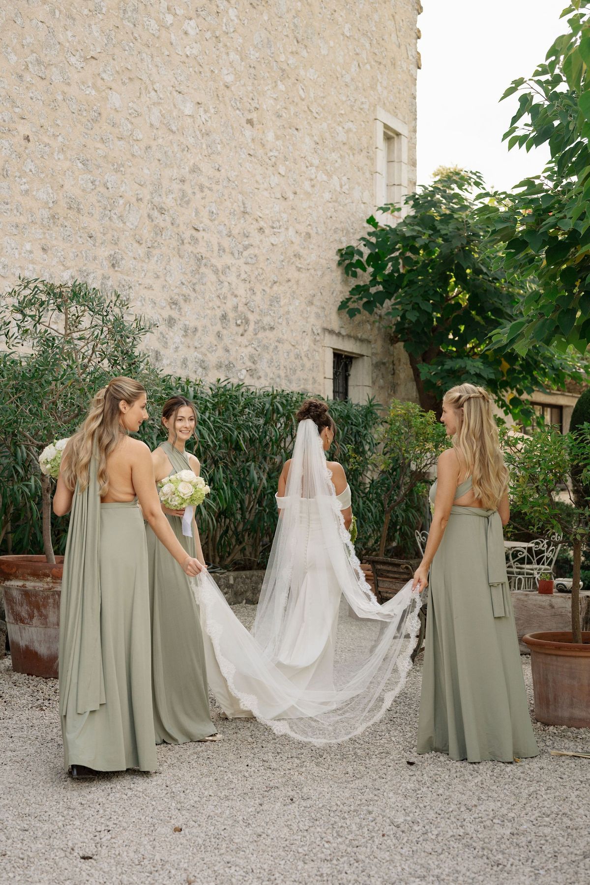 Bridesmaids in sage gowns extending bride's cathedral veil at chateau