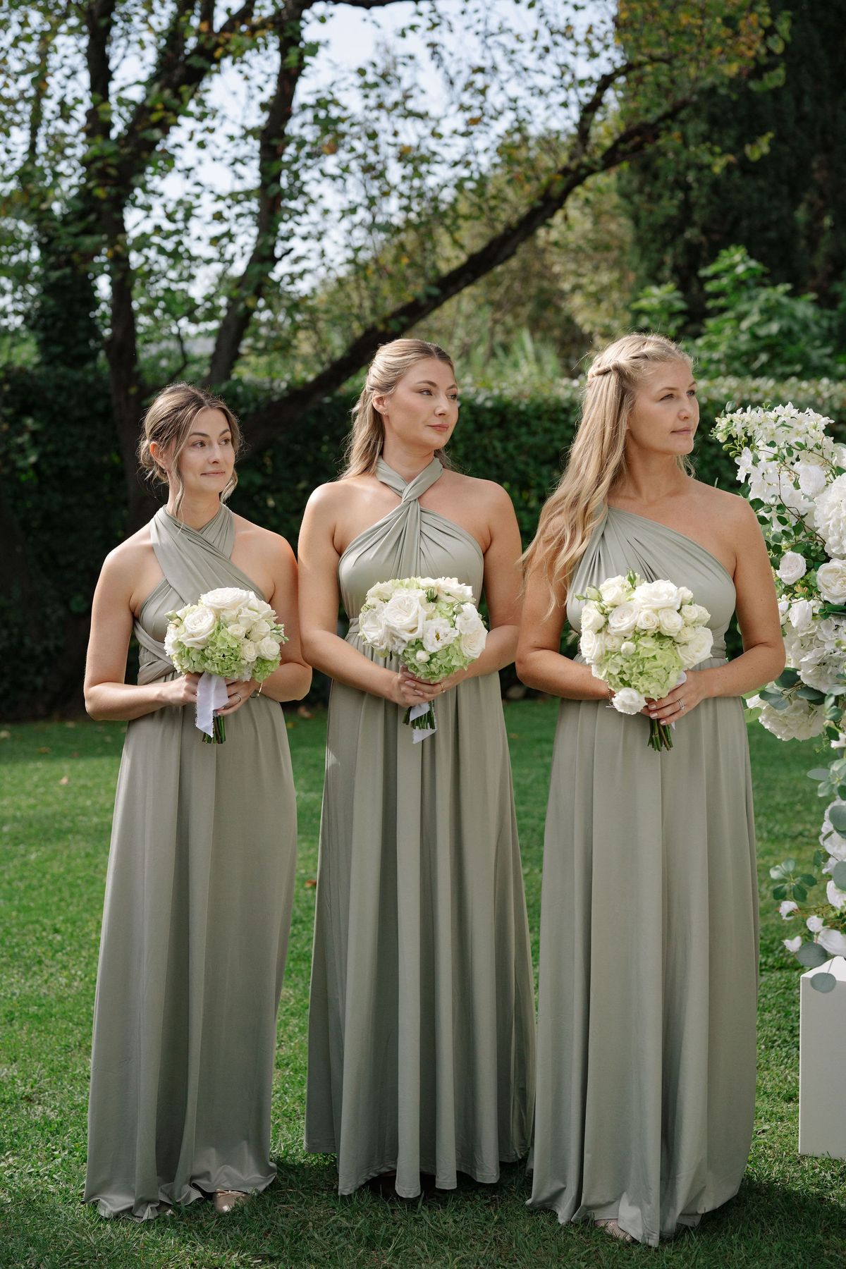 Three bridesmaids in sage gowns holding white bouquets at ceremony