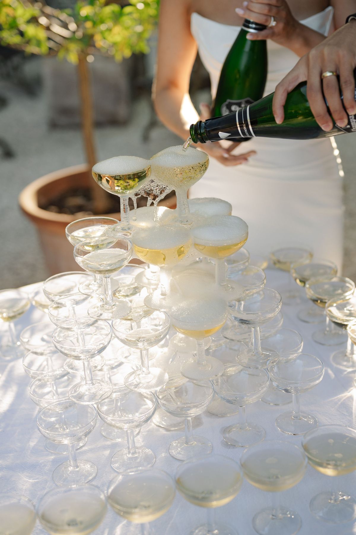 Bride pouring champagne over coupe glass tower at outdoor reception