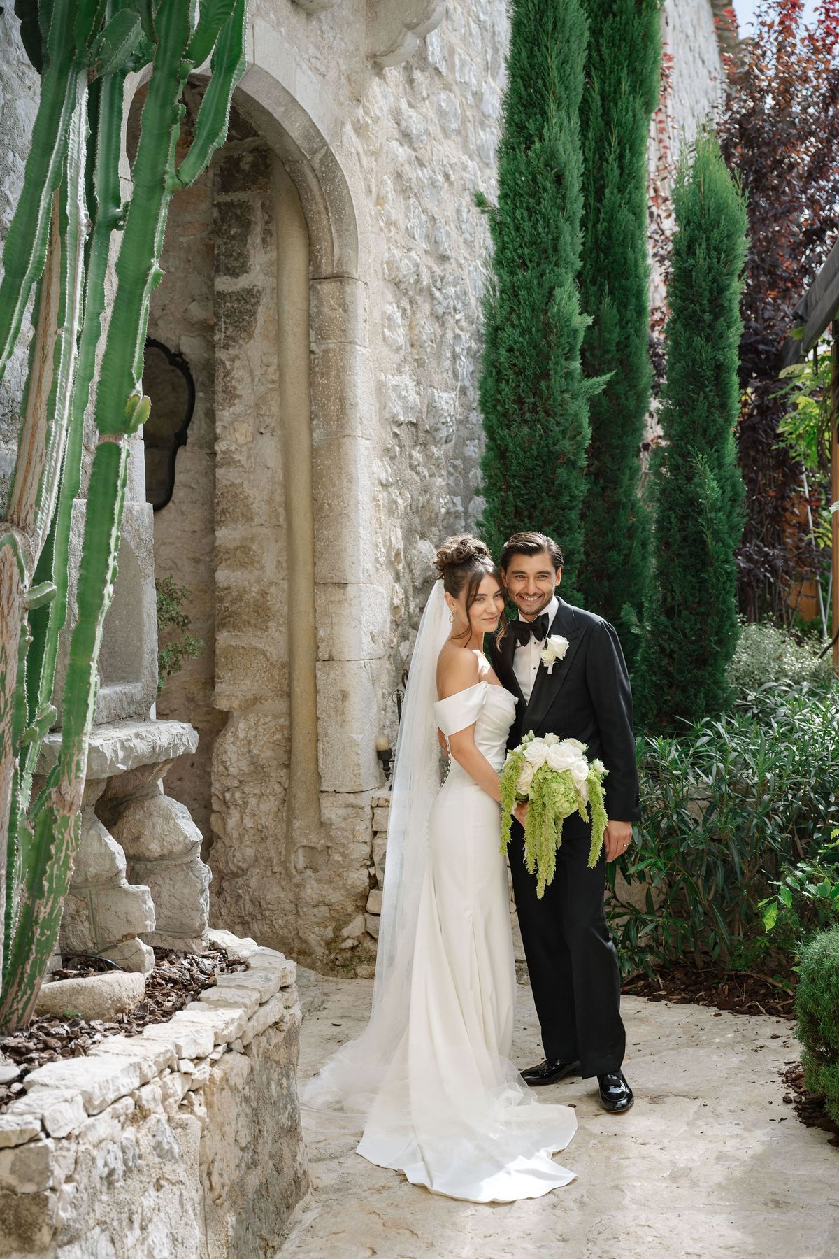 Bride and groom standing cheek to cheek beside stone arch in garden
