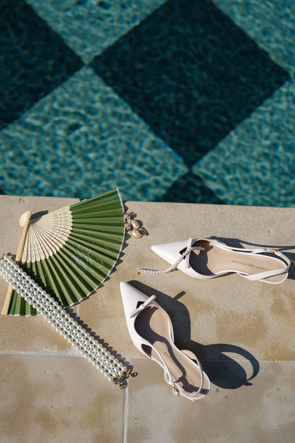 Flat lay of bridal shoes, fan, and pearl jewellery beside checkerboard pool