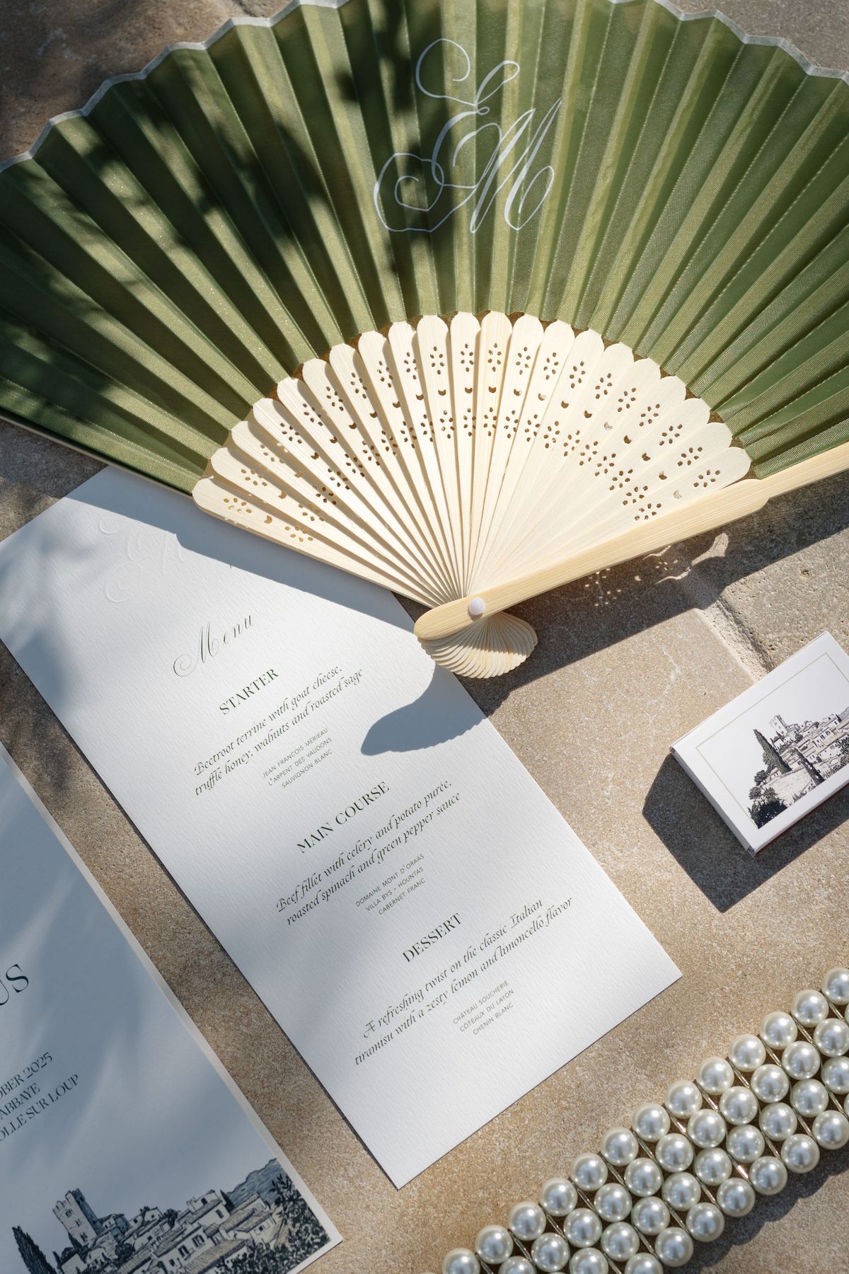Flat lay of sage paper fan, printed menu, matchbook, and pearl bracelet
