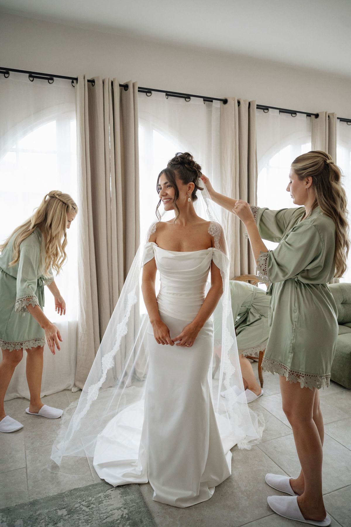 Bridesmaids fitting cathedral veil onto bride in sunlit getting-ready room
