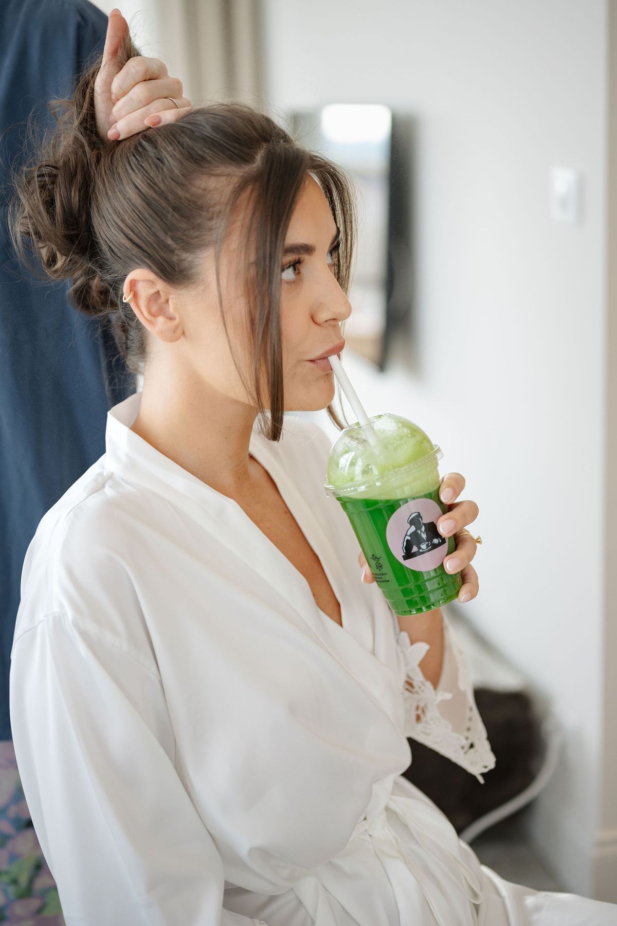 Bride sipping green slushie while stylist adjusts her hair in robe