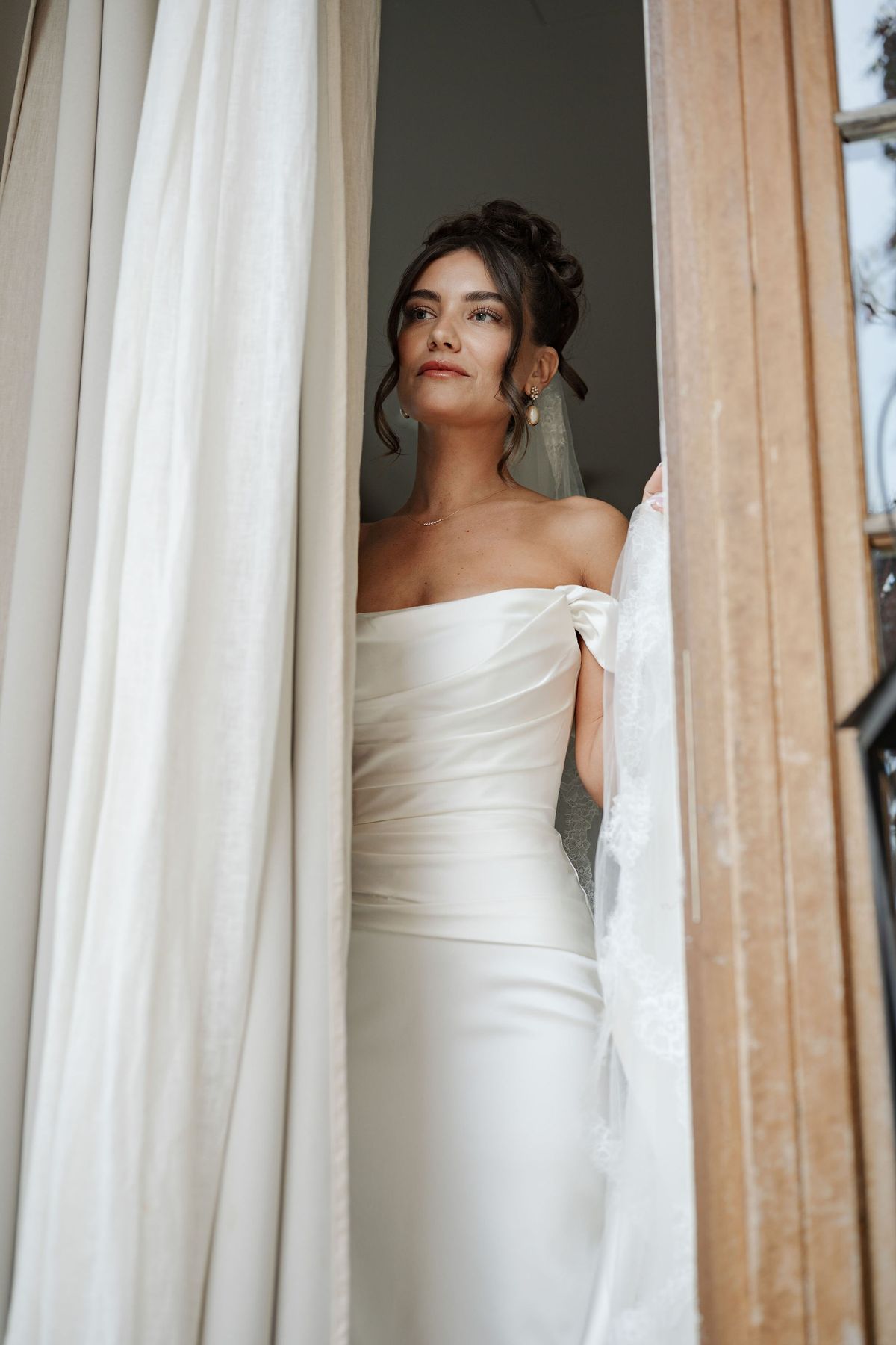 Bride gazing out doorway between linen curtains in satin gown