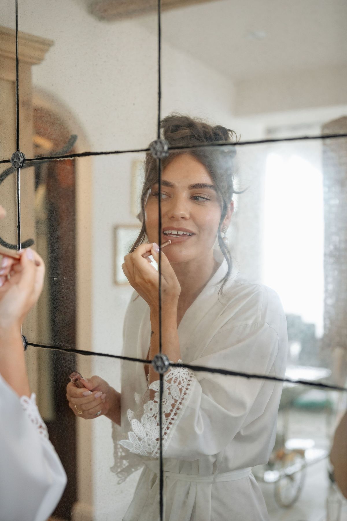 Bride applying lipstick in leaded mirror while wearing white lace robe