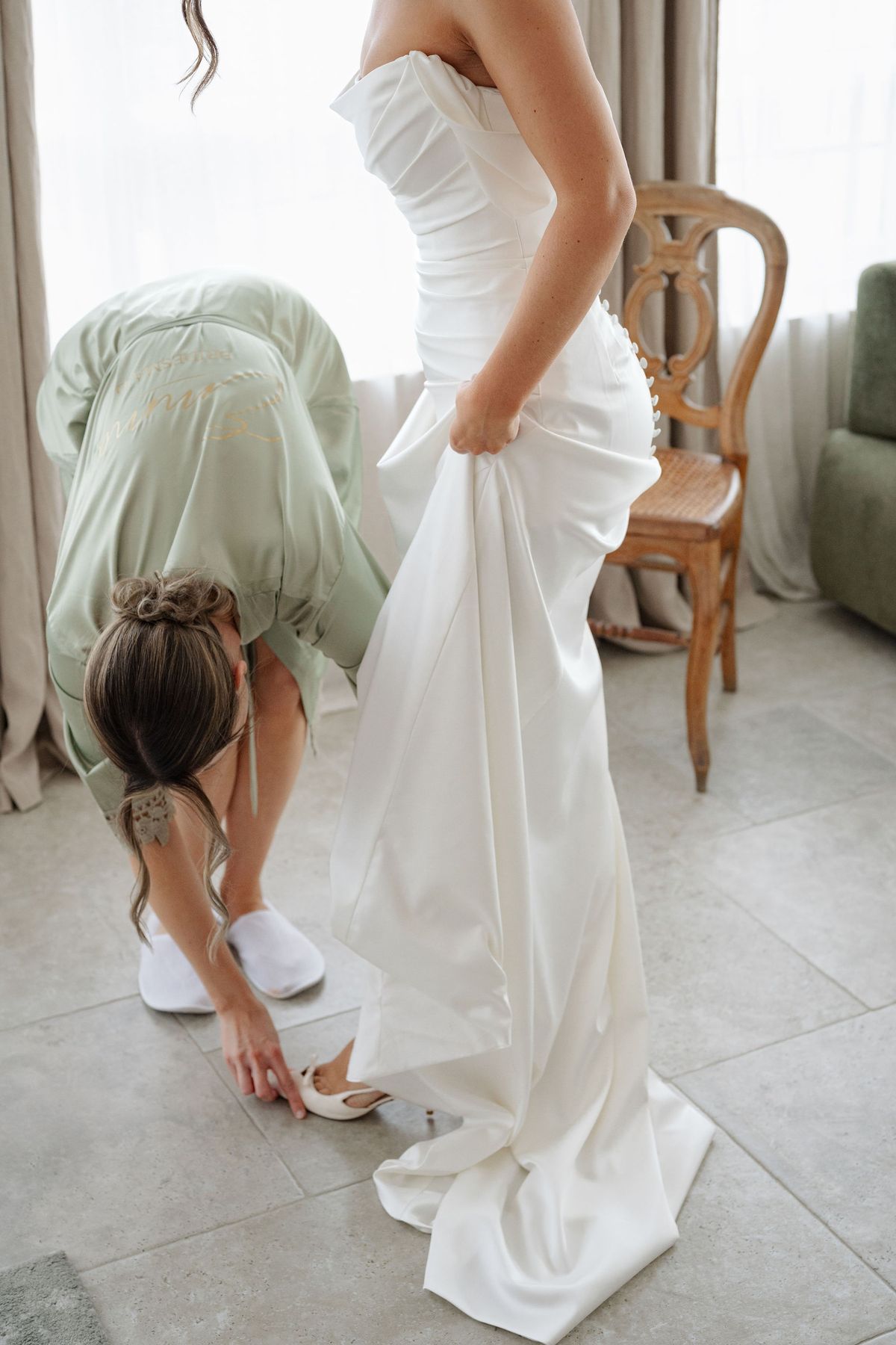 Bridesmaid in sage robe helping bride into her pointed white shoes