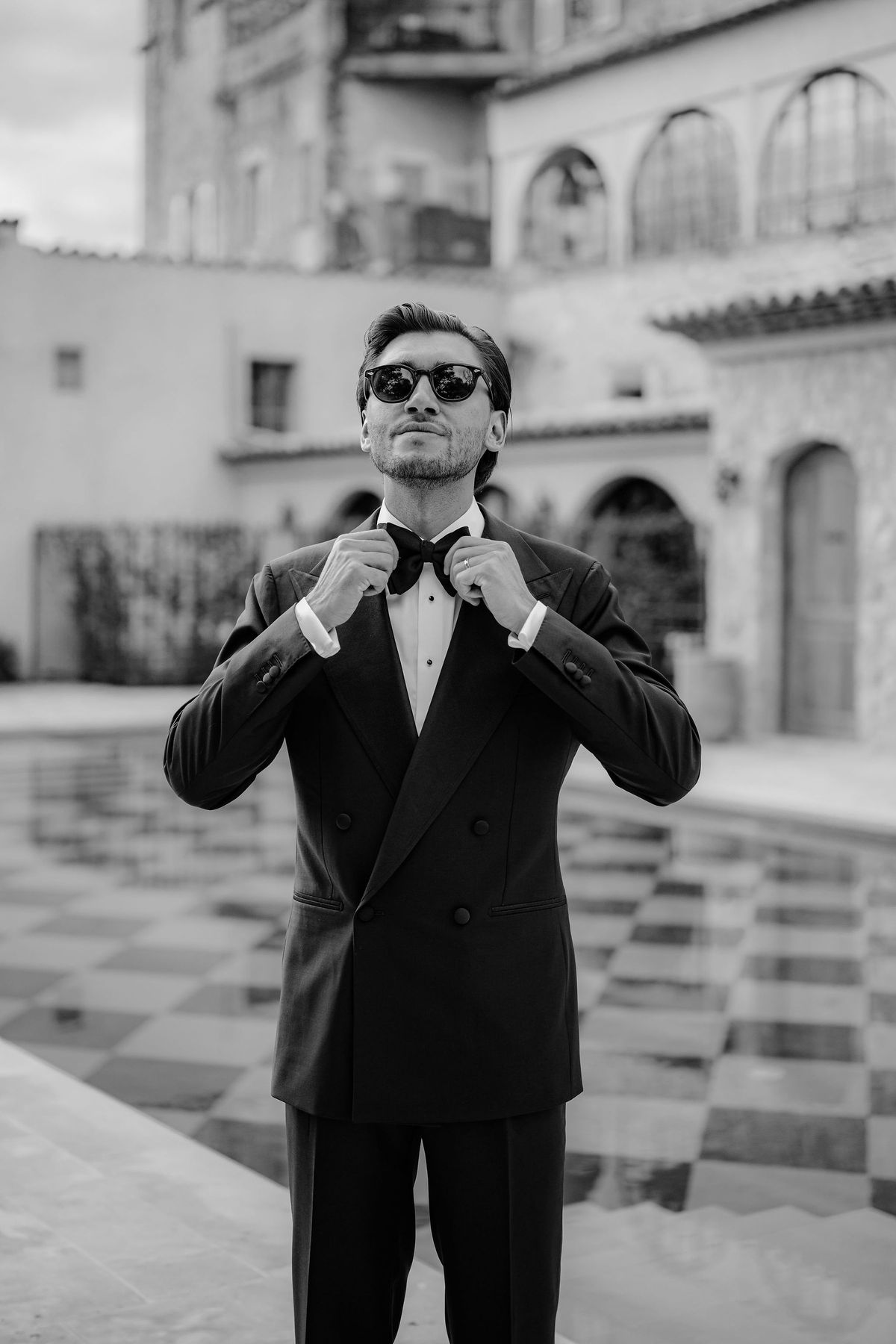 Groom in tuxedo adjusting bow tie beside checkerboard pool in black and white