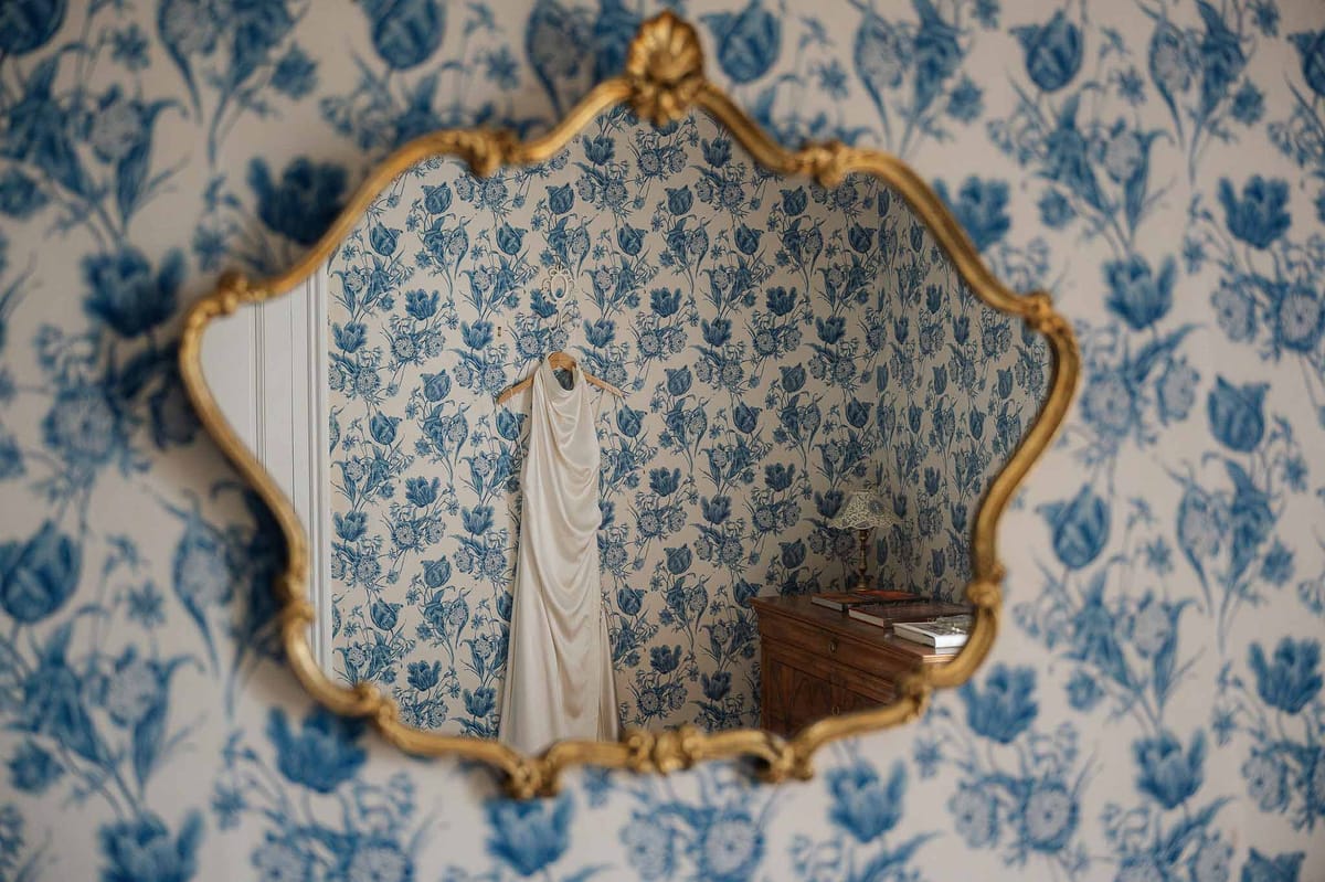 Ivory satin wedding dress reflected in gold-framed mirror on blue and white toile wallpaper in French interior