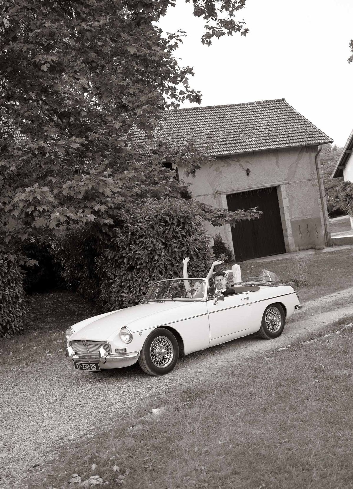 Black and white wide shot of couple departing in vintage white MGB convertible on gravel drive