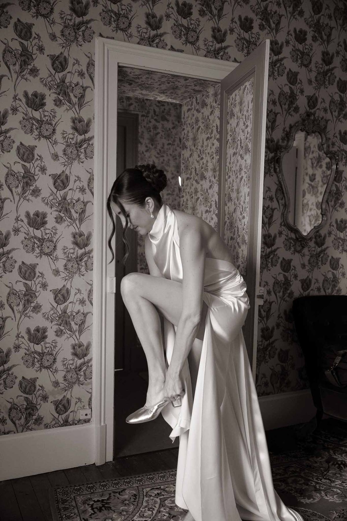 Bride in satin halter gown bends to put on shoe in floral-wallpapered doorway in B&W