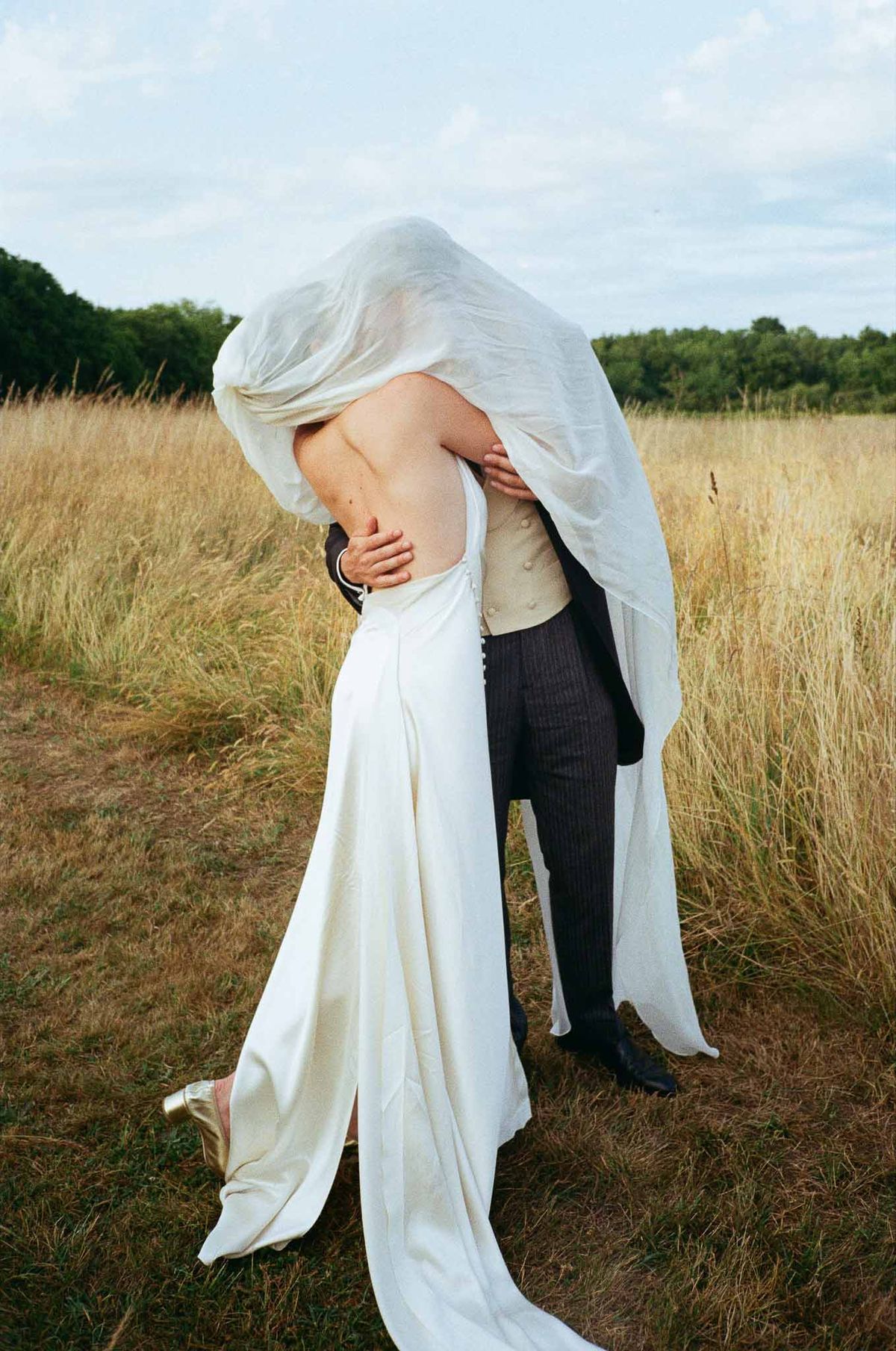 Couple kissing under tulle veil in golden grass field with bride in low-back satin gown and gold heels