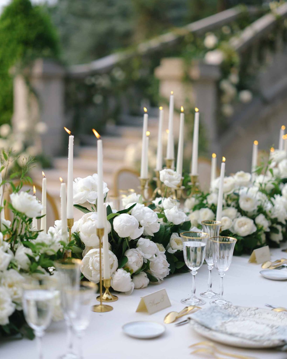 White peony runner with gold candelabras, gold flatware, and crystal glassware under greenery-draped pergola