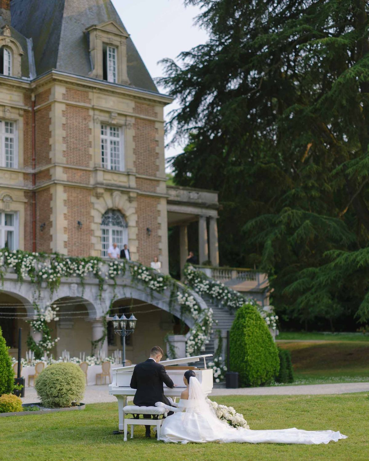 Bride and groom at white grand piano on lawn with rose-draped double staircase behind at chateau
