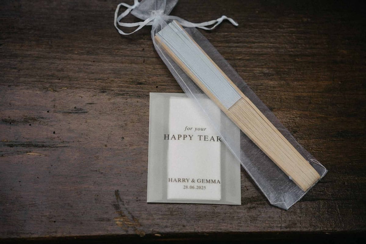 Wedding favour hand fan in organza bag with personalised happy tears card on dark wood surface
