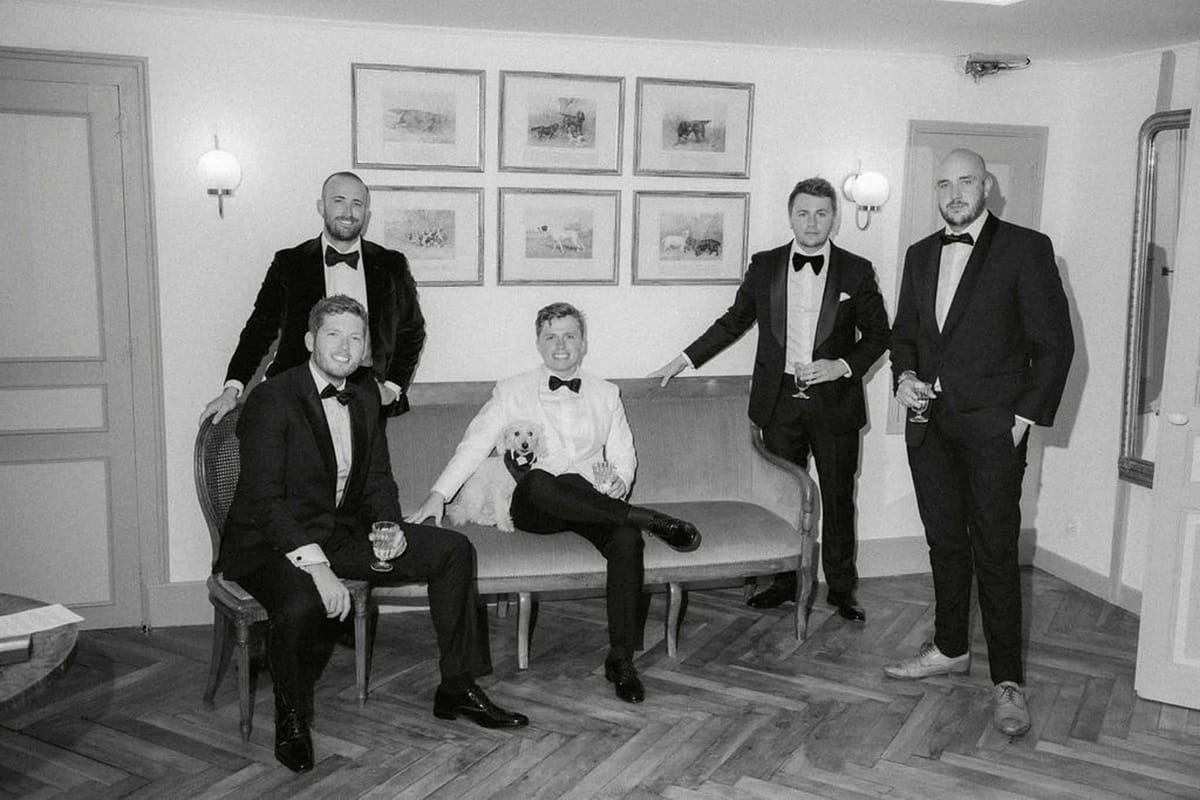 Groom in white dinner jacket with small dog and four groomsmen in tuxedos on settee in salon in B&W