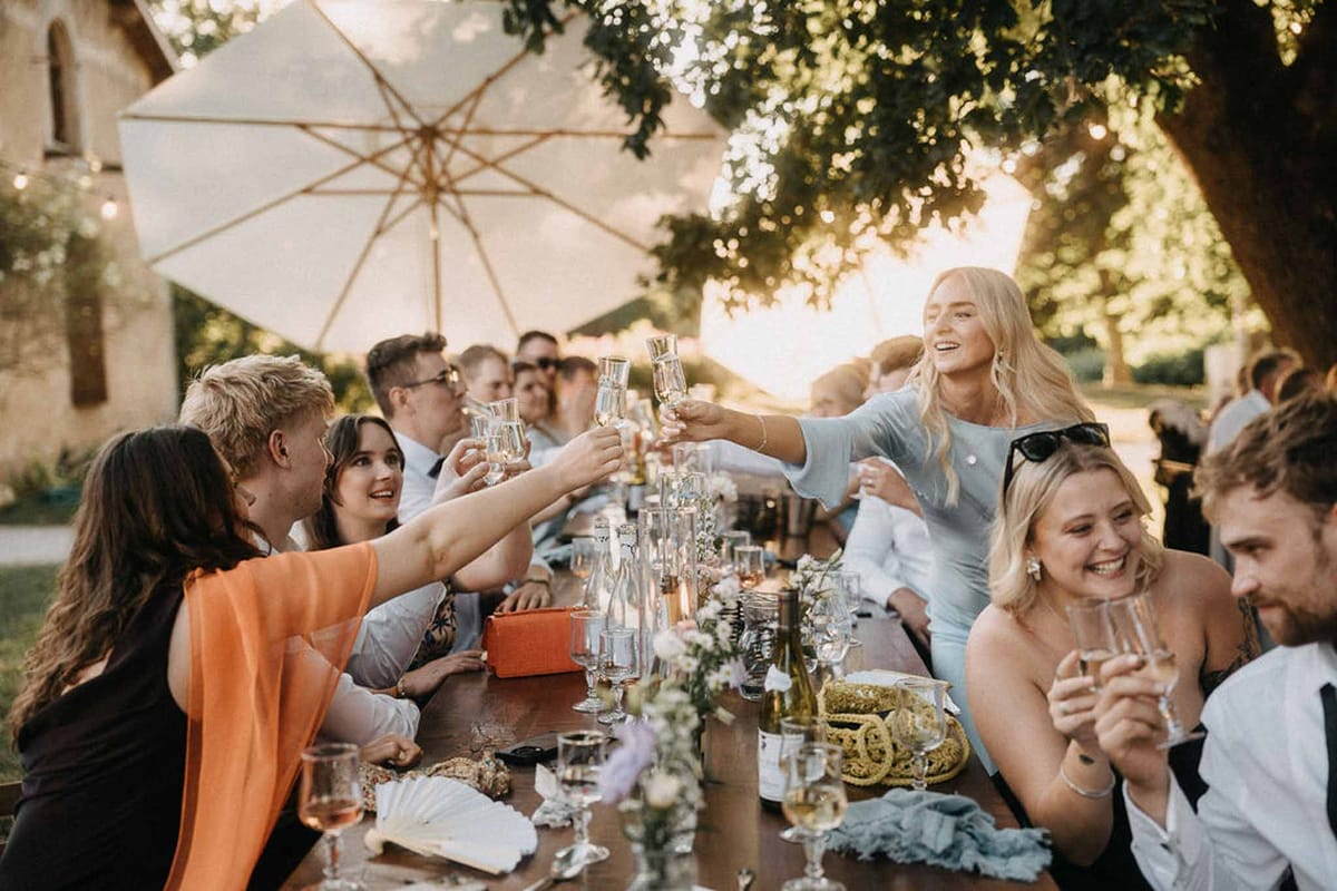 15 guests raising champagne flutes at farm table under fairy lights at golden hour by stone building