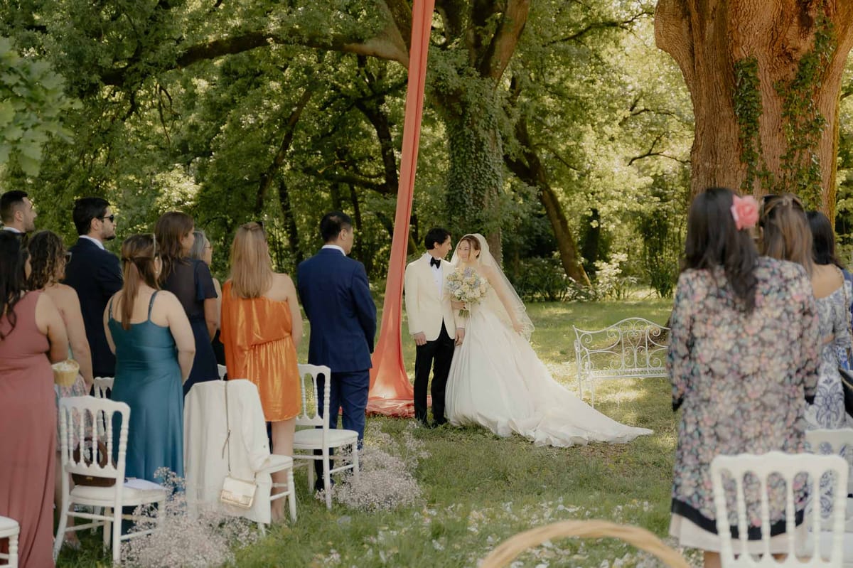 Outdoor woodland ceremony with couple at altar under trees, white Chiavari chairs, and baby's breath aisle