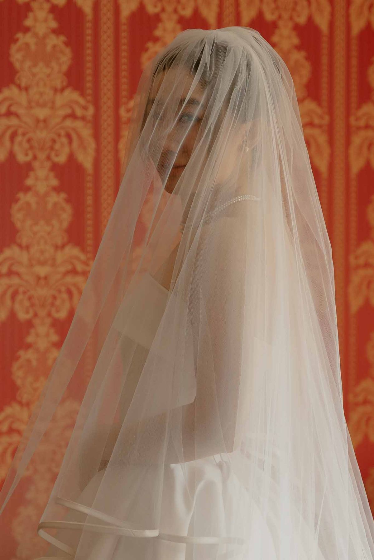 Bride draped in ivory tulle veil against red and gold damask wall with pearl-trimmed neckline visible beneath