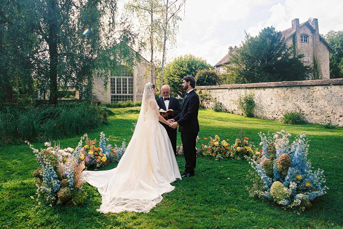 Bride and groom hold hands at garden ceremony with wildflower aisle of blue delphiniums and orange ranunculus