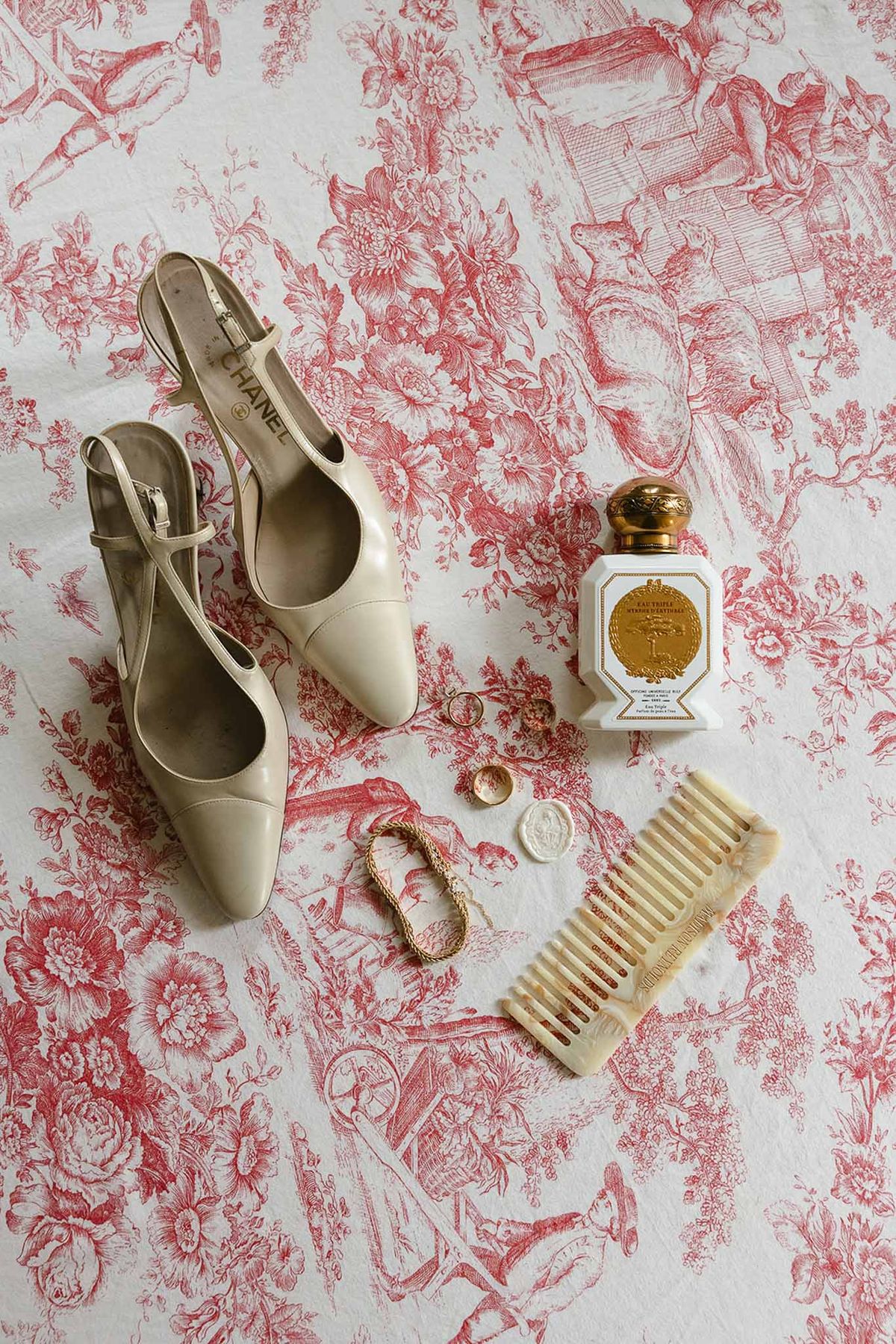 Bridal flat lay with ivory Chanel slingbacks, gold perfume bottle, and horn comb on red toile fabric