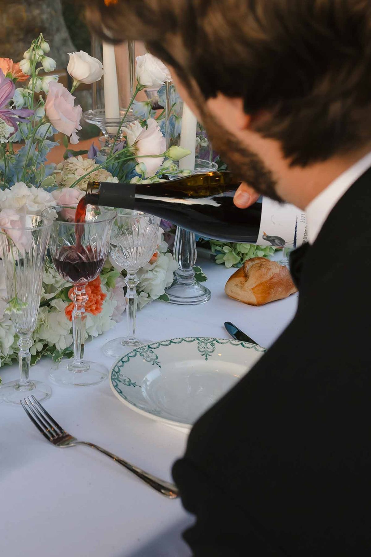 Guest pouring red wine beside blush rose and blue delphinium centerpiece with crystal stemware and china