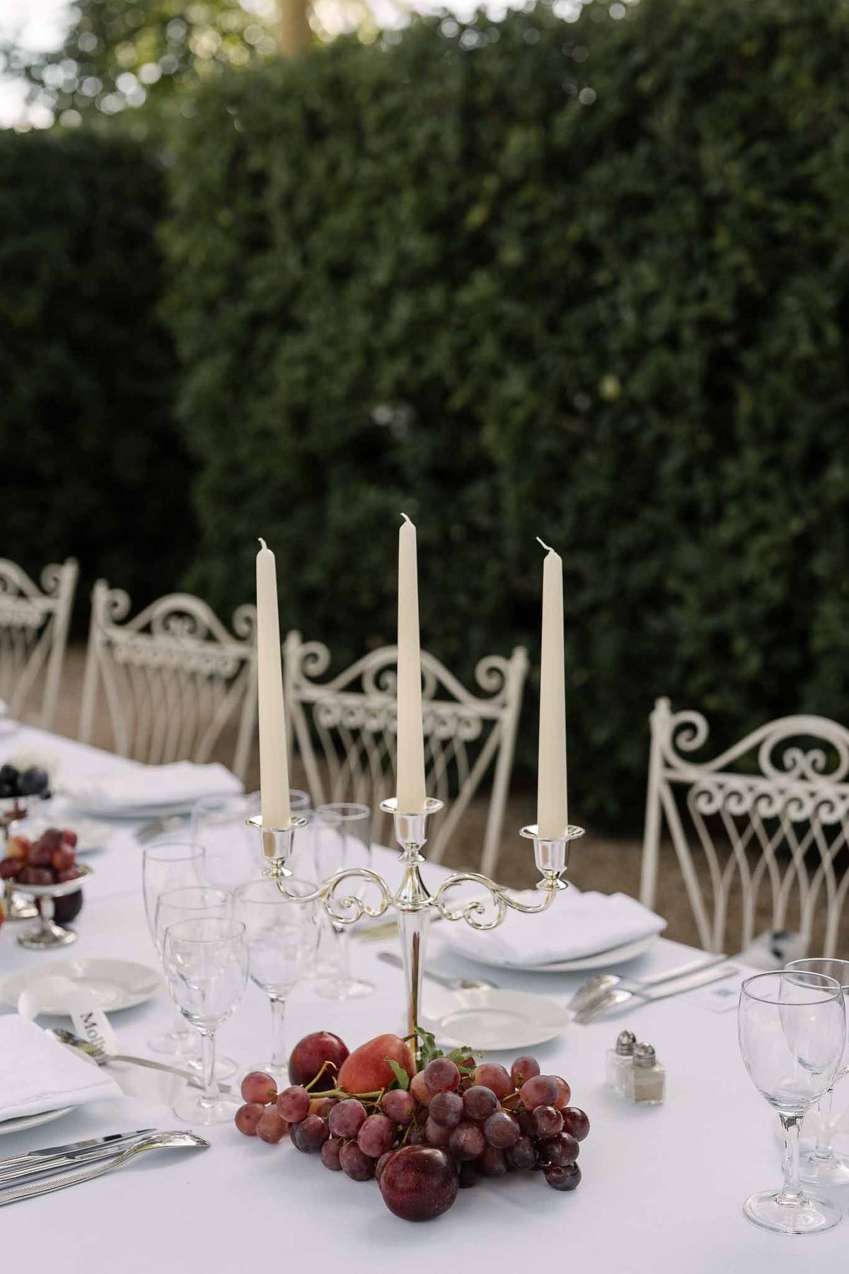 Silver three-arm candelabra with ivory tapers, red grapes, and stone fruit on white garden table