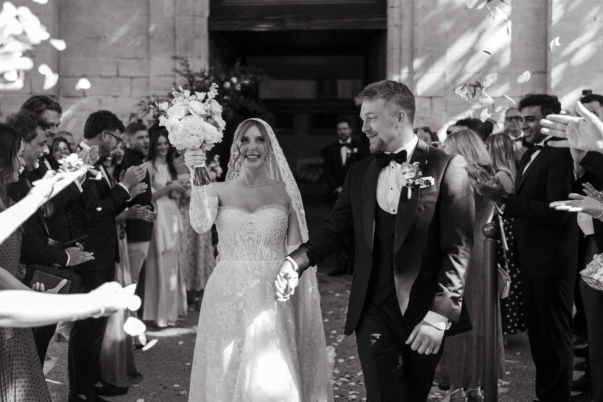 Black and white church exit with bride raising peony bouquet as guests throw petals under stone arch