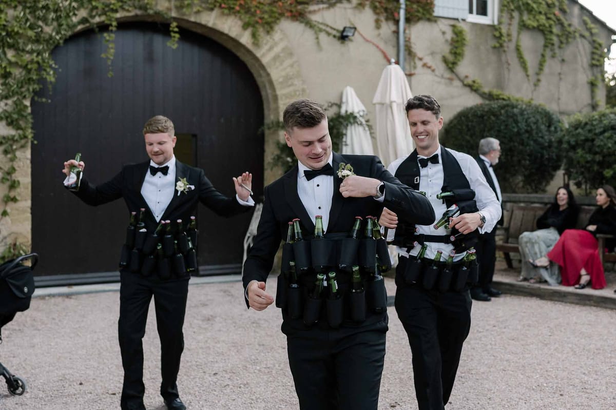 Three groomsmen in tuxedos wearing novelty beer holster vests distributing drinks in chateau courtyard