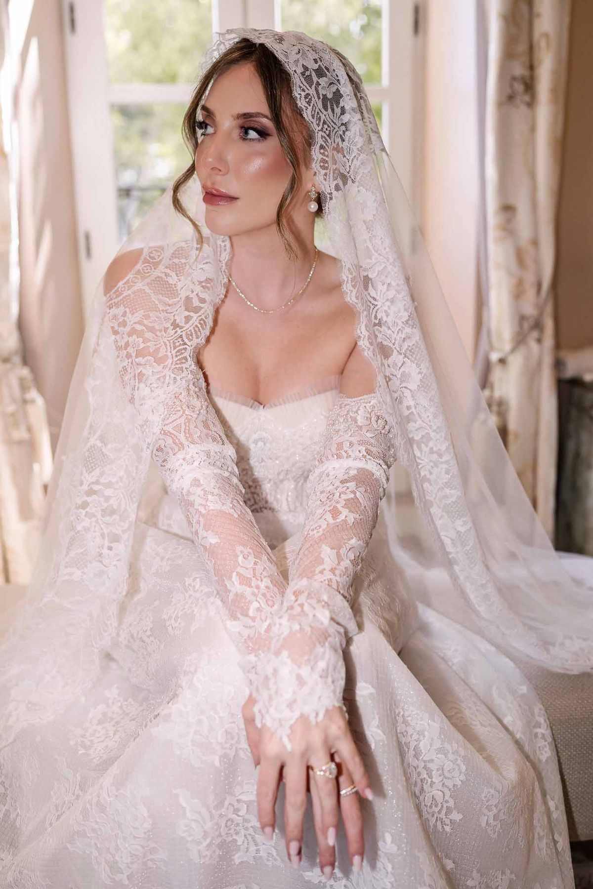Bride in ivory lace gown with mantilla veil, pearl earrings, and smoky eye makeup near chateau window