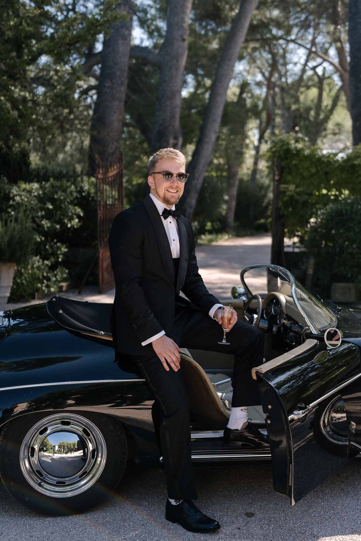 Groom in black tuxedo and bow tie leaning against vintage black convertible holding champagne flute on tree-lined driveway