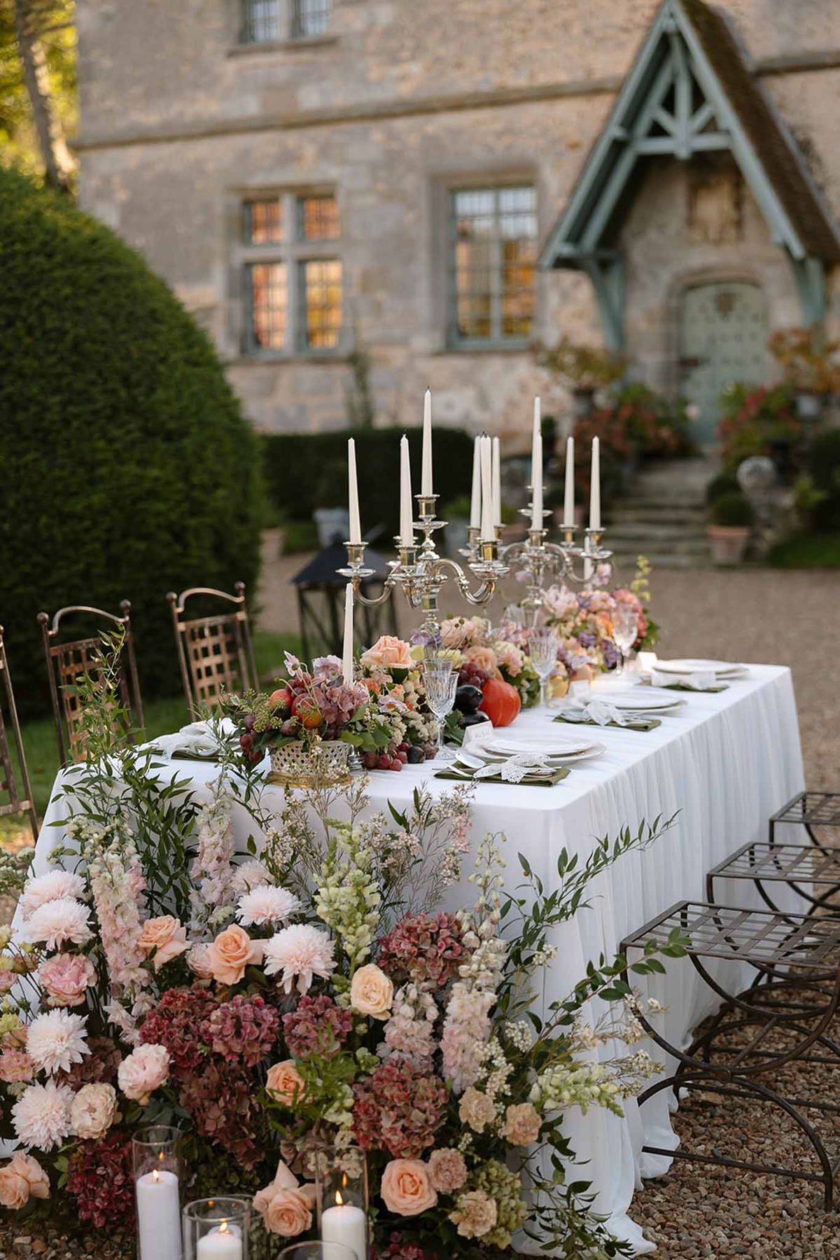 Long table with blush roses, mauve hydrangeas, silver candelabras, and hurricane candles before stone manor