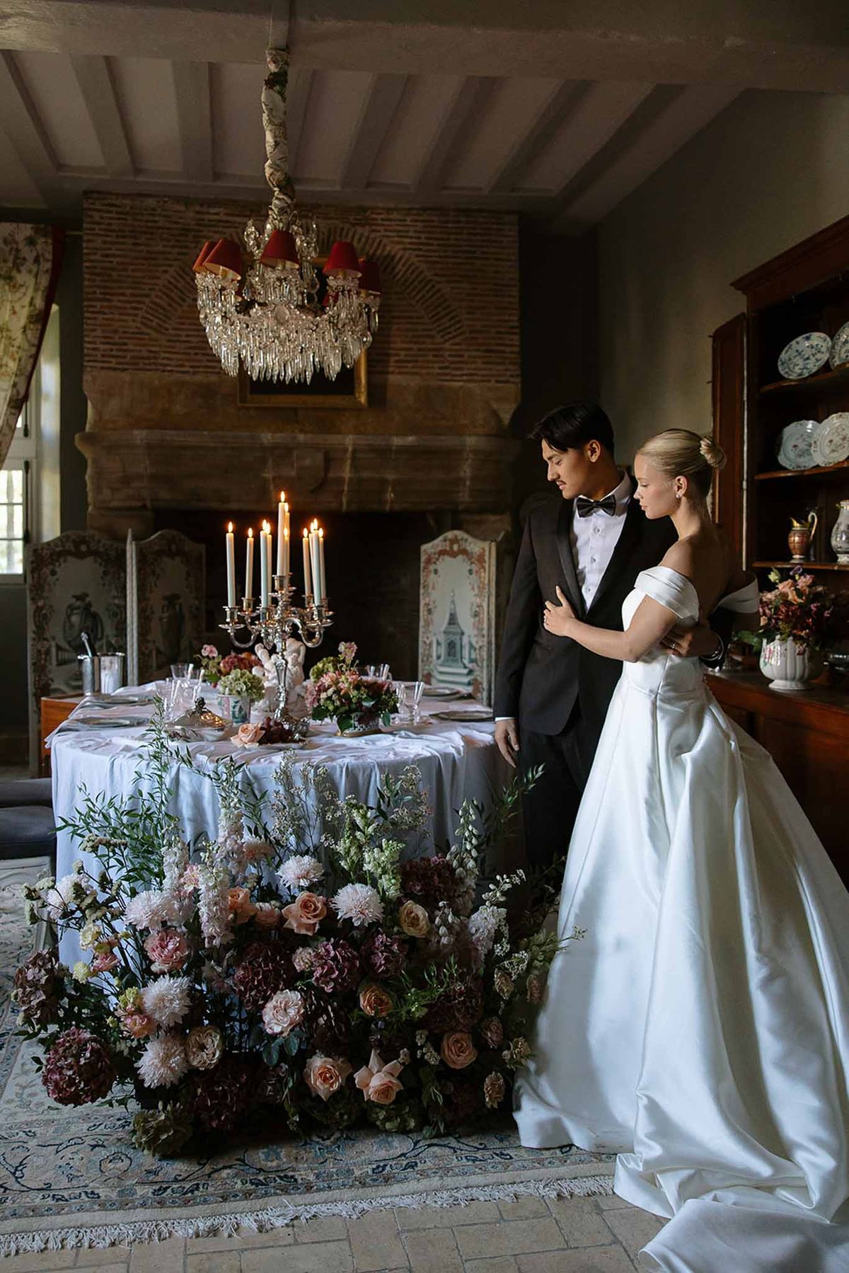 Couple beside sweetheart table with cascading burgundy dahlias and blush roses in moody chateau salon