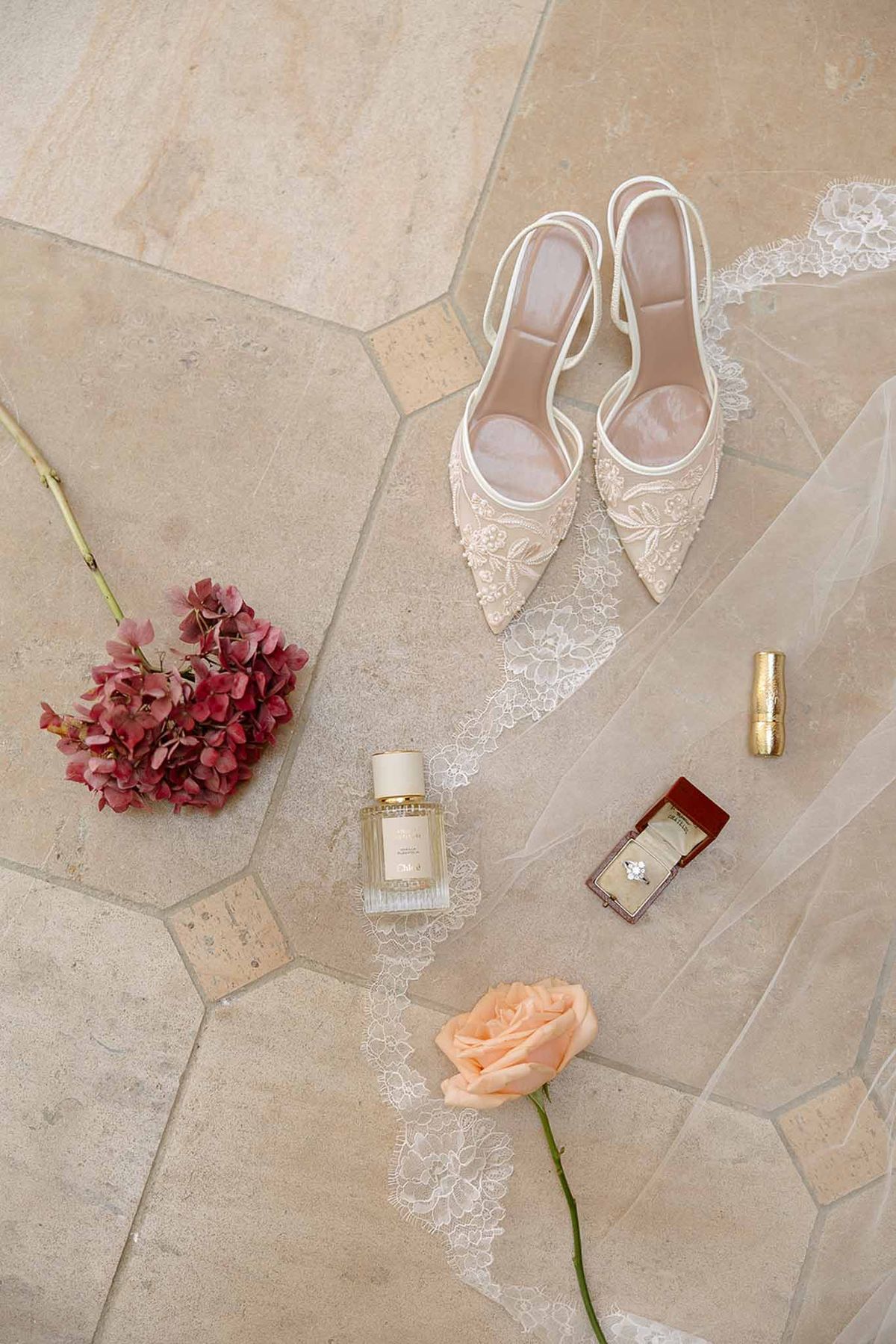Bridal flat lay with ivory floral heels, lace veil, diamond ring in red velvet box, perfume, and peach rose