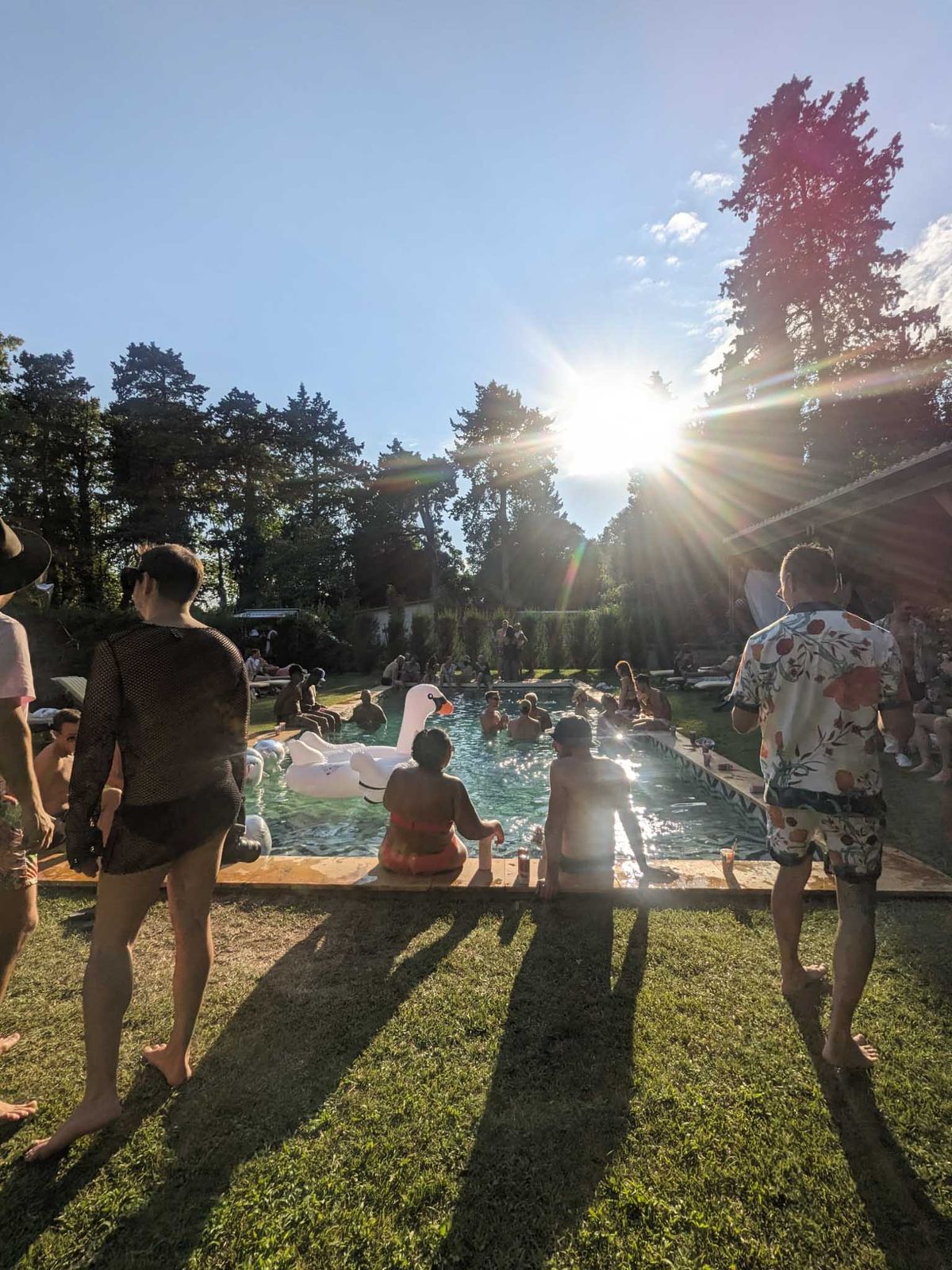 Pool party at golden hour with guests swimming and sitting poolside beside white inflatable swan on estate lawn