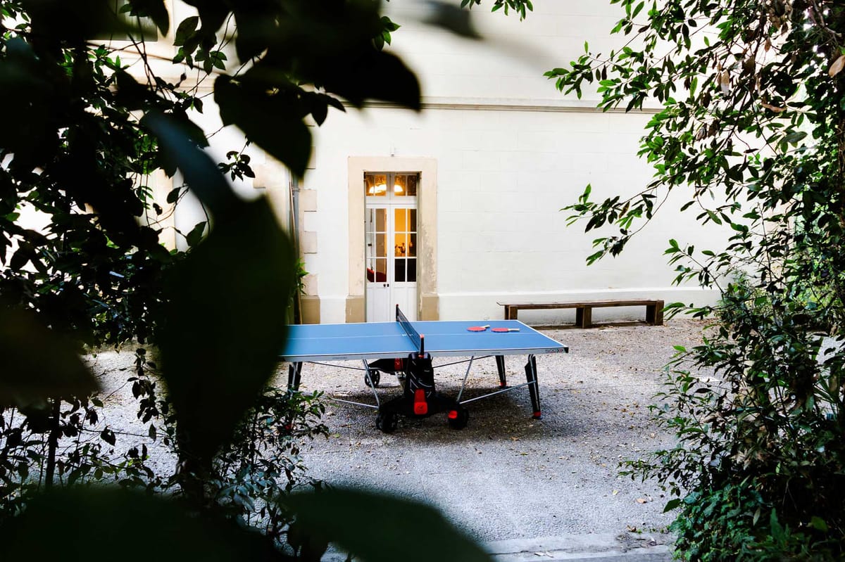 Courtyard table tennis table with red paddles on gravel beside glowing chateau doorway at evening