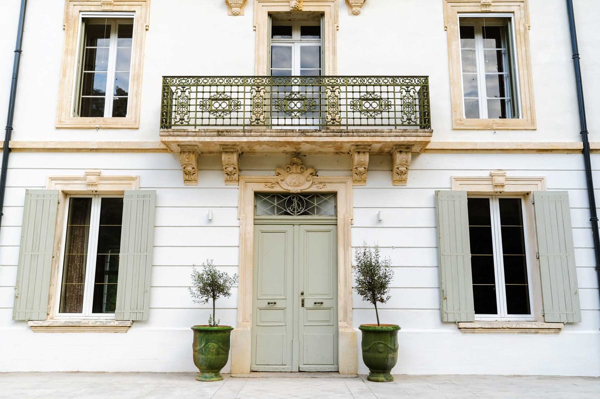 White bastide entrance with sage green doors wrought-iron transom stone cartouche and olive trees in Anduze urns