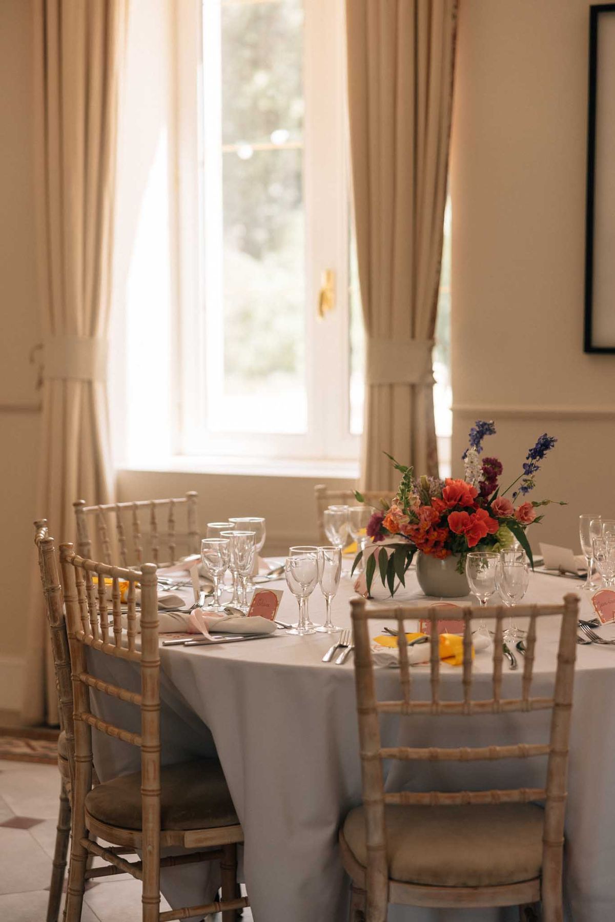 Round reception table with grey-blue linen, coral ranunculus and purple delphinium centerpiece, blush napkins, and chiavar...