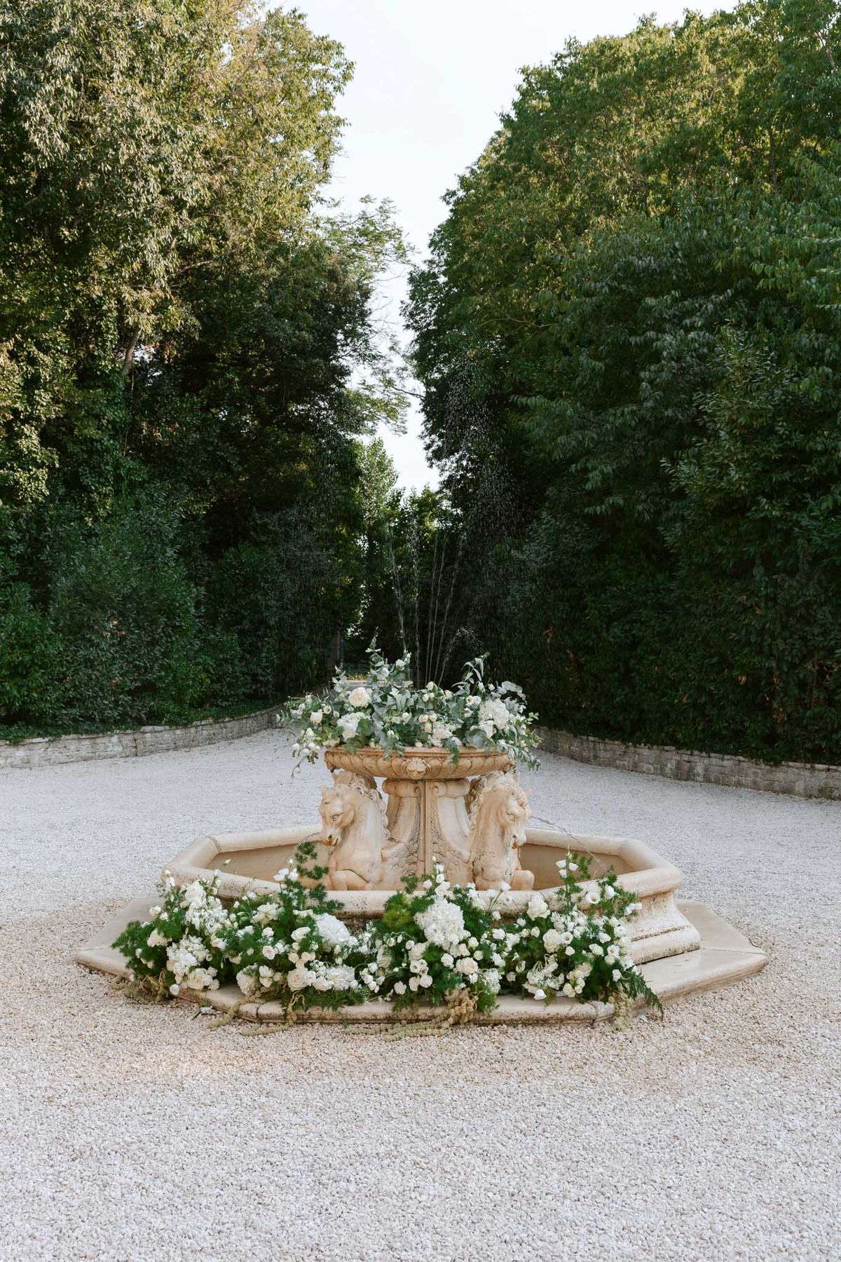 Stone horse fountain decorated with ivory roses, white ranunculus, and trailing greenery on gravel courtyard