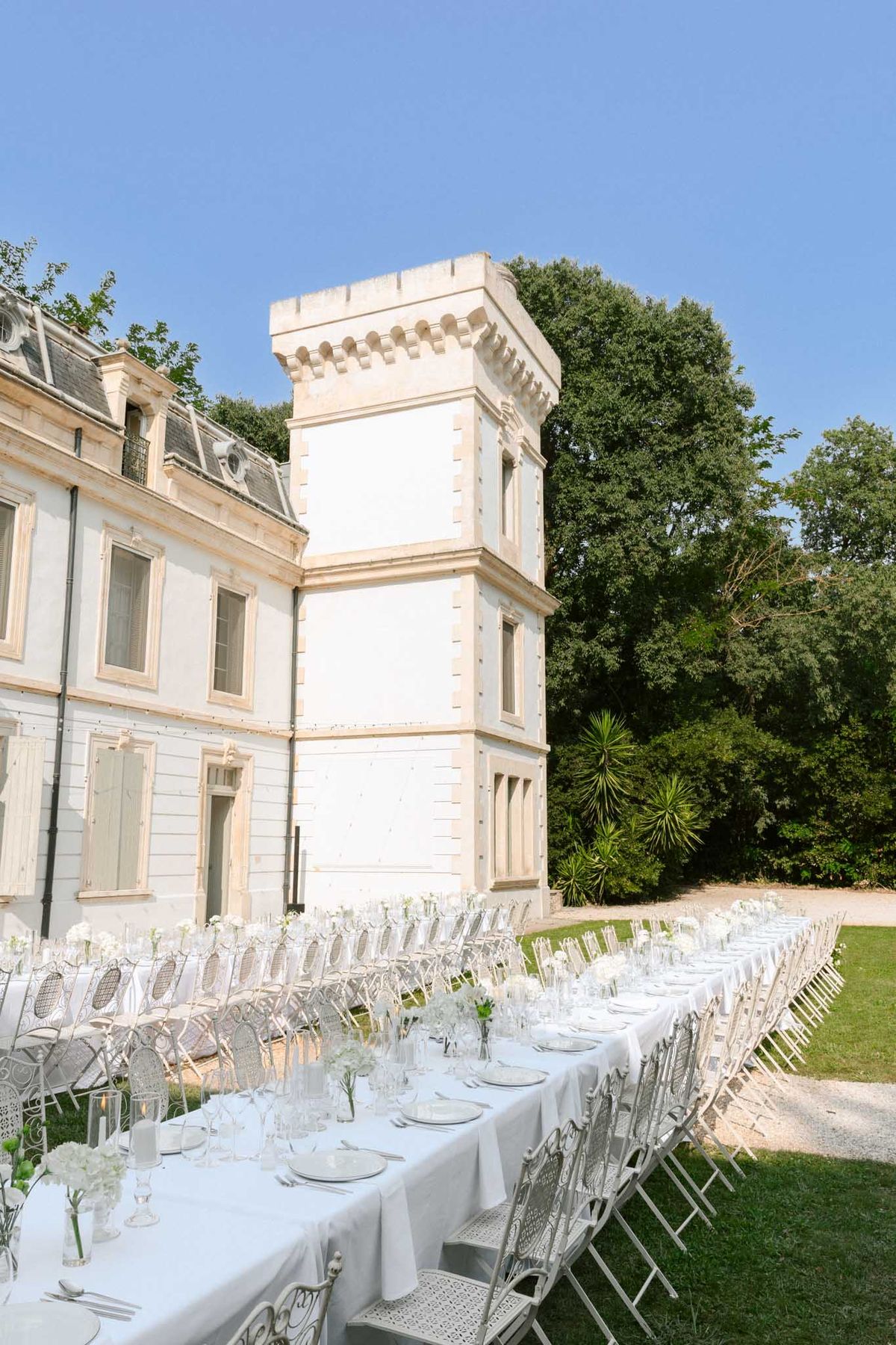 Long white linen tables with hydrangea bud vases and metal garden chairs before stone chateau with square tower