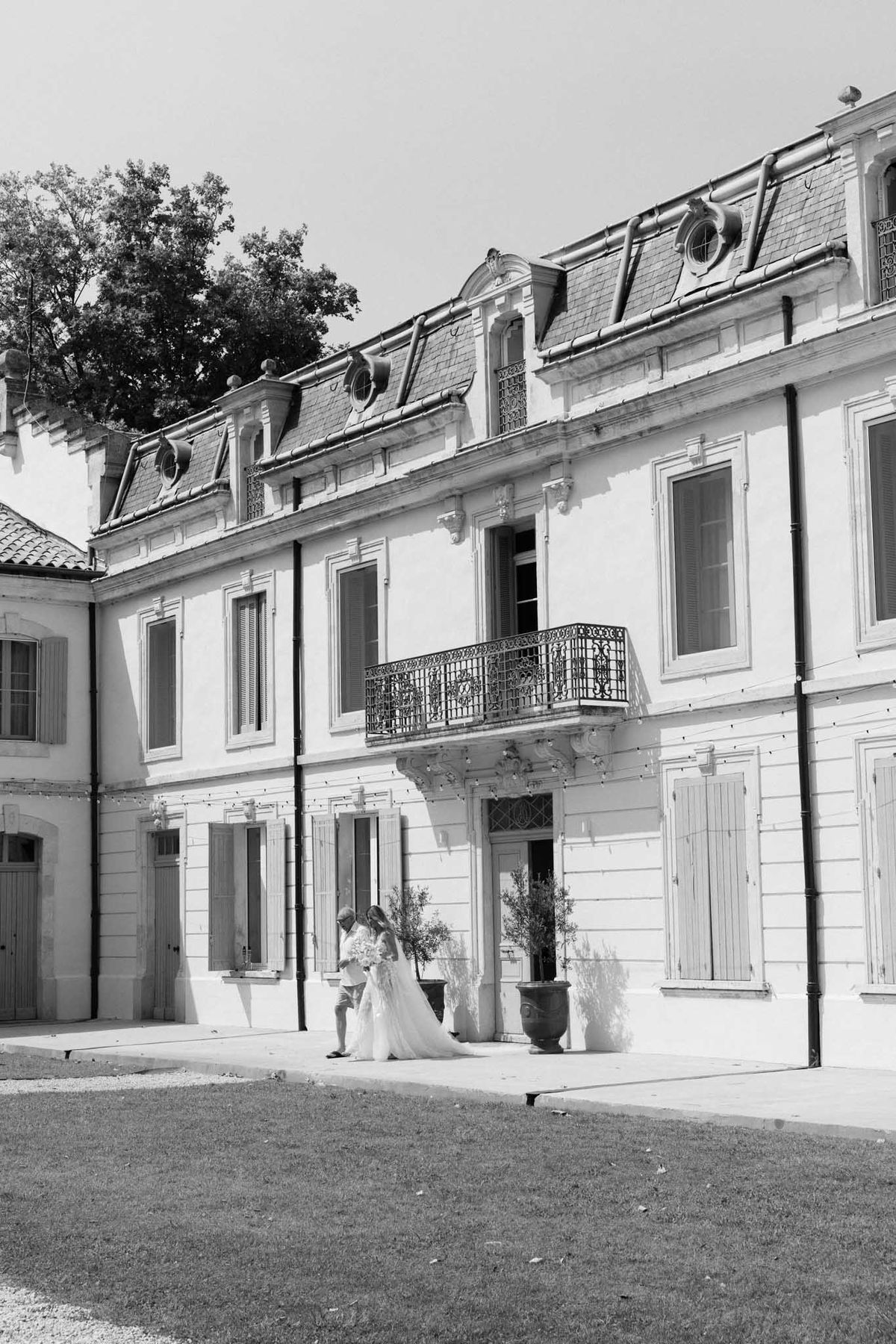 Black and white wide shot of bride walking along classical French chateau exterior with mansard roof
